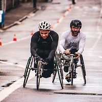 Kolorowa fotografia. Trwa uliczny wyścig osób niepełnosprawnych. Dwóch zawodników ściga się na specjalnych wózkach